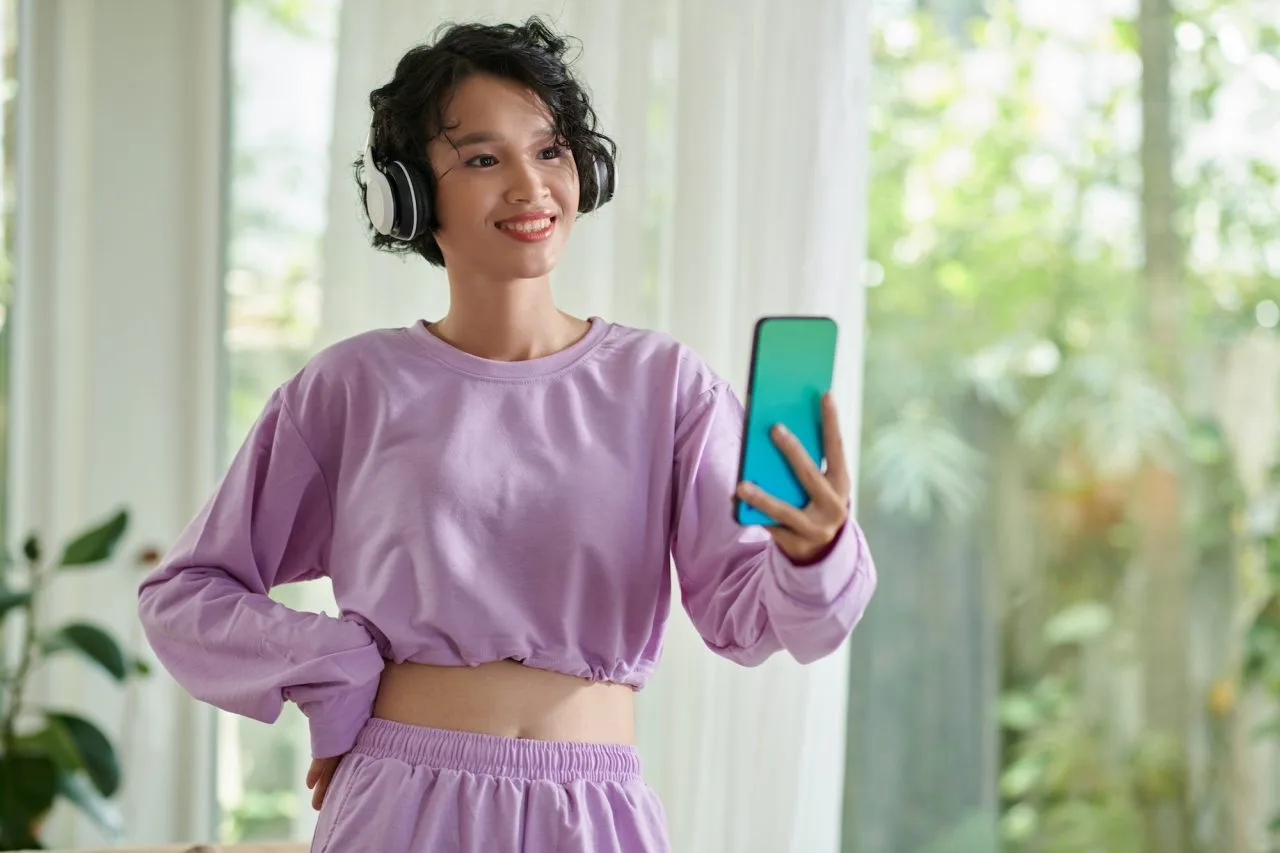 Uma jovem de cabelo curto e moletom roxo usa fones de ouvido e sorri enquanto olha para seu smartphone.